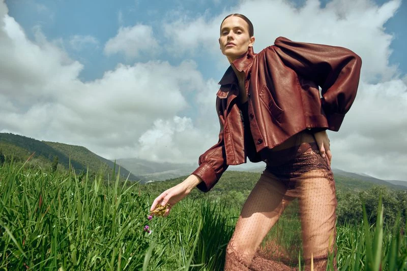 A fashion photo showcases a model in a brown leather jacket and sheer pants in a tall grassy field with a mountain background.