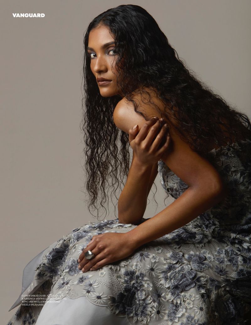 Portrait of a model with long dark curly hair wearing a floral dress while seated against a neutral backdrop, posing for a beauty and fashion shoot.