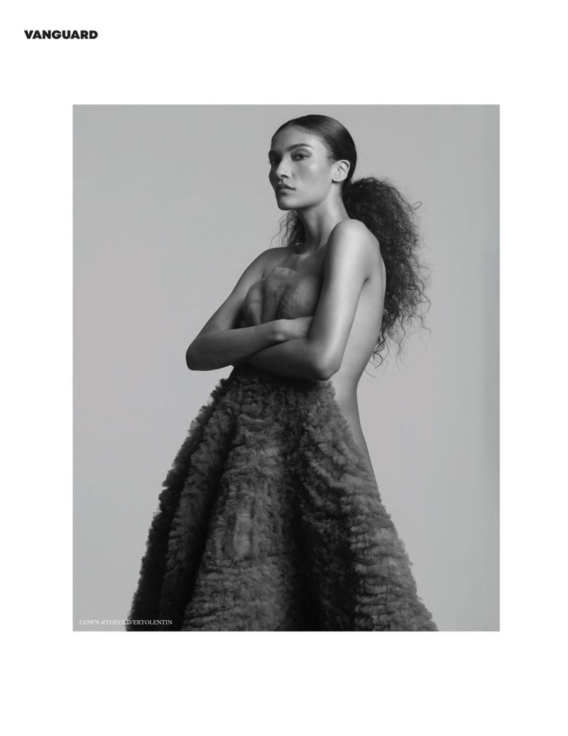 Black and white portrait of a woman in a large, textured dress posing in front of a neutral backdrop with arms crossed and ponytail.