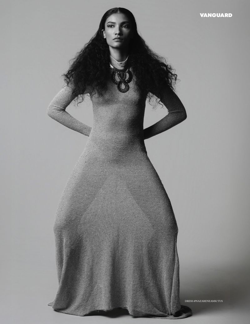 Black and white fashion shot of a woman in a long dress posing in front of a grey background, wearing a necklace.