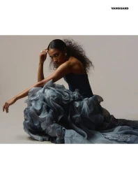 Model seated on grey floor wearing voluminous ruffled dark blue gown against a plain light background