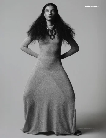 Model with long dark curly hair wearing a long fitted dress and statement necklace against a grey studio background