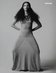 Model with long dark curly hair wearing a long fitted dress and statement necklace against a grey studio background