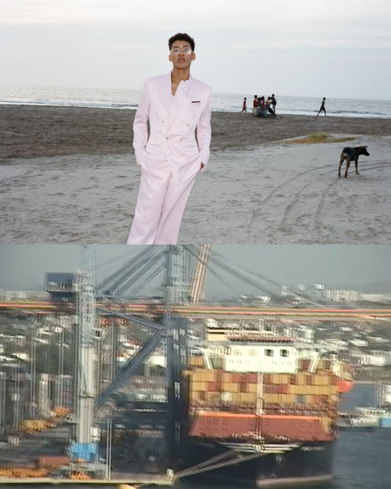 Man in a pale pink suit standing on a sandy beach near the ocean with people in the background, with a view of ships and cranes.