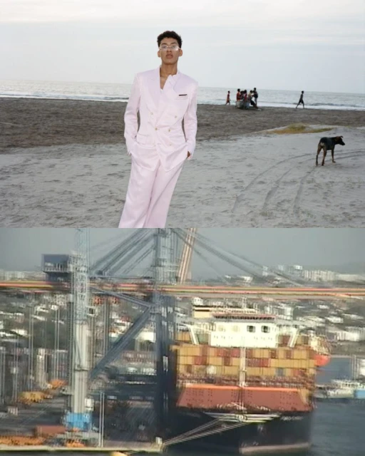 Man in a pale pink suit standing on a sandy beach near the ocean with people in the background, with a view of ships and cranes.