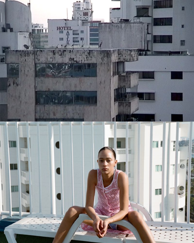 Adult woman in pink dress sits on white lounge chair, set against cityscape backdrop in bright daylight.