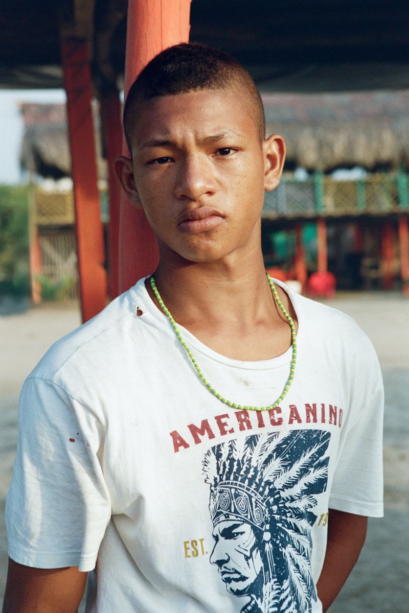 Portrait of a young man with a serious expression, wearing a graphic t-shirt and green beaded necklace outdoors under a wooden structure.