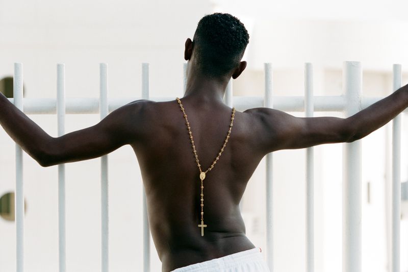 Man with dark skin and short hair, shot from behind, wearing a gold rosary and white shorts, leaning against a white metal railing.