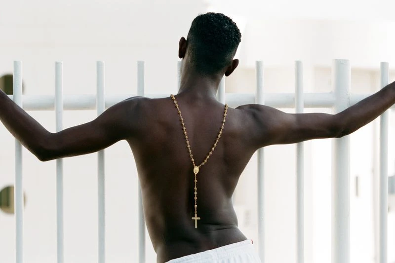 Man with dark skin and short hair, shot from behind, wearing a gold rosary and white shorts, leaning against a white metal railing.