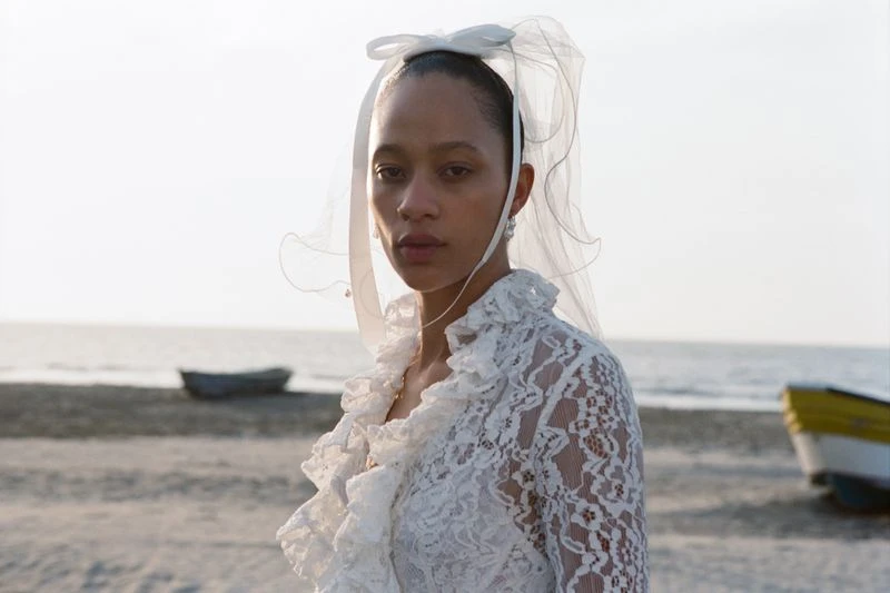 A woman wearing a white lace dress and veil stands on a beach with boats in the background on a bright day, captured in a medium shot.