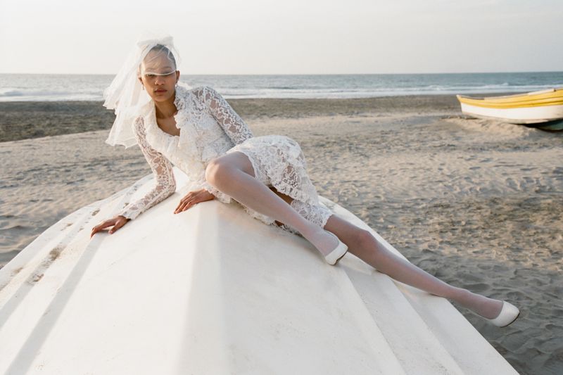 Woman in a lace wedding dress and veil posing on a white boat at a beach, with the sea in the background on a sunny day.