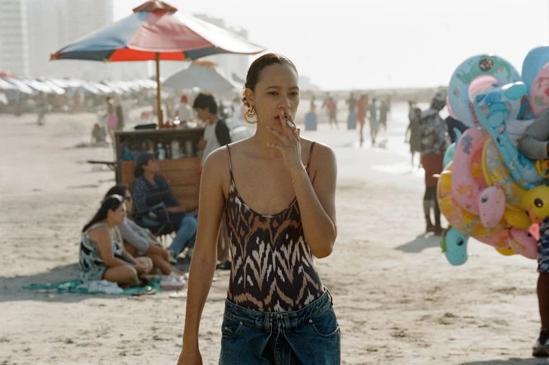 Woman in patterned swimsuit and denim skirt smokes on a sandy beach; beachgoers and colorful floats fill the sunny background.