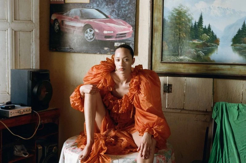 A fair-skinned adult woman in an orange ruffle dress poses indoors, seated on a floral tablecloth with vintage decor and natural lighting.