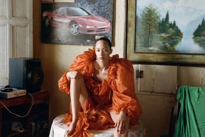 A fair-skinned adult woman in an orange ruffle dress poses indoors, seated on a floral tablecloth with vintage decor and natural lighting.