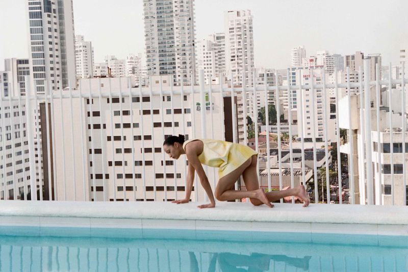Woman in a yellow dress crawls on the edge of a rooftop pool with a city skyline backdrop on a bright sunny day.
