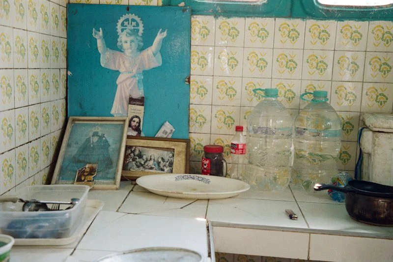 Indoor shot features religious art, plastic water bottles, and kitchenware on a white tile counter against patterned tiles.