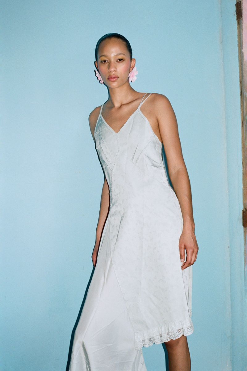 A young woman with fair skin and pink flower earrings poses in a vintage white slip dress against a light blue wall.