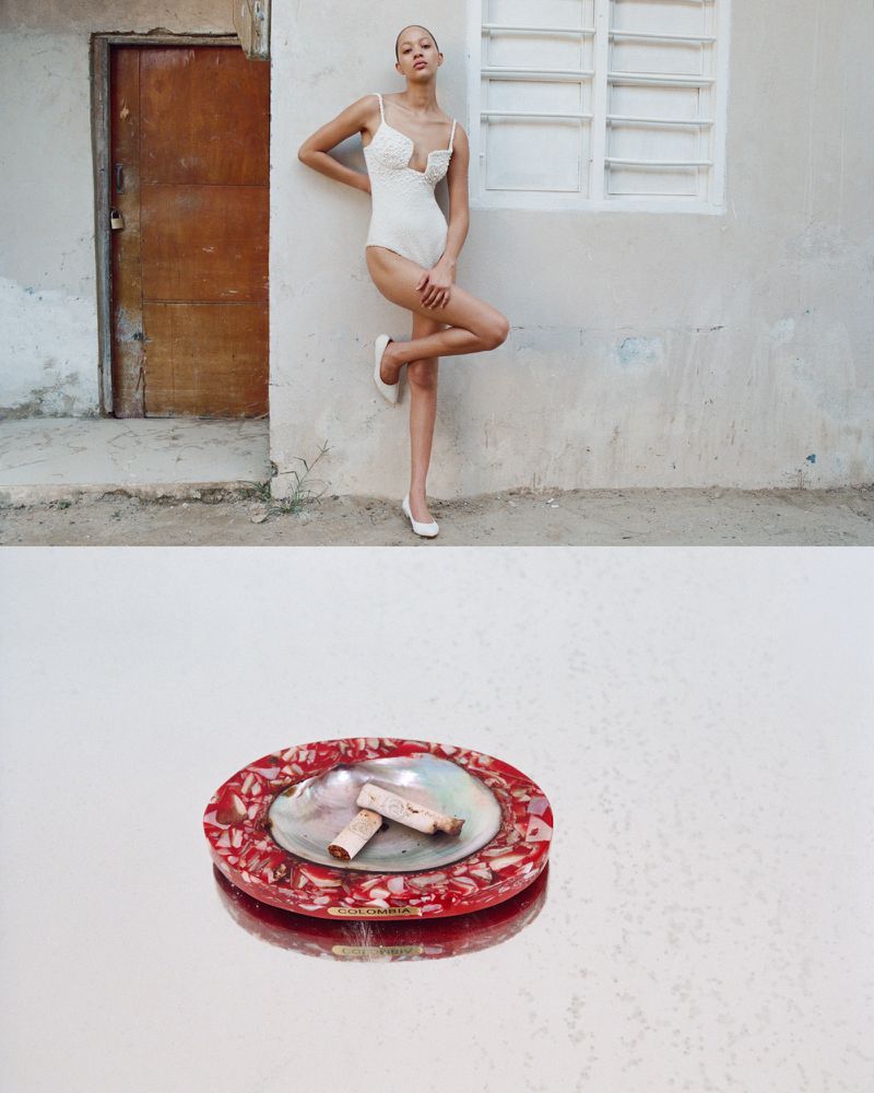A slender model poses in a white bodysuit and ballet flats against a textured wall, with a decorative ashtray in the foreground.