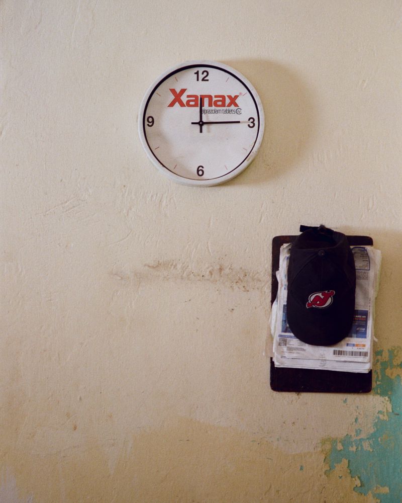 Clock with the word 'Xanax' on the face is mounted on a stained wall, beneath it hangs a baseball cap and papers attached to the wall.