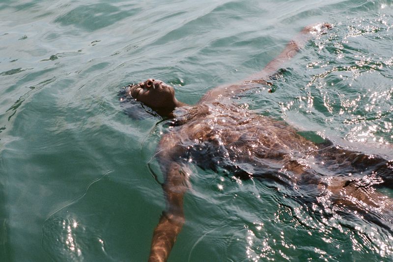 Person floats on back in shimmering turquoise water with arms outstretched, face visible in sunlight, creating a tranquil water scene.
