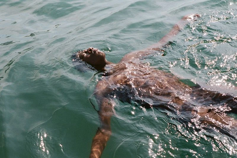 Person floats on back in shimmering turquoise water with arms outstretched, face visible in sunlight, creating a tranquil water scene.