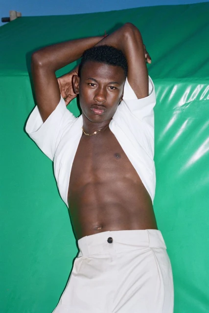 An athletic Black male model poses with arms raised in a photo studio. He's wearing white pants and a white top. The background is a green screen.