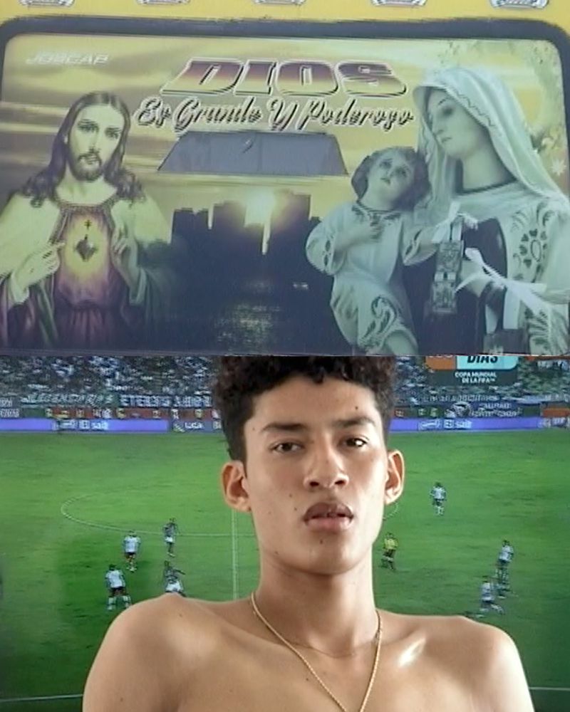 Young adult shirtless man poses in front of a television screen showing a soccer game and a religious illustration.
