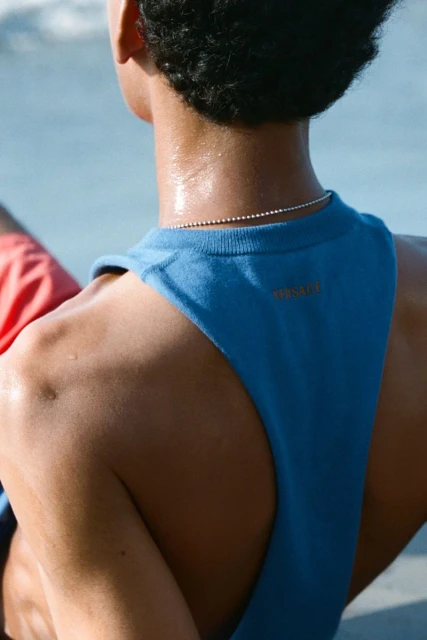 Back view of an athletic man in a blue tank top with sweat, wearing a silver chain against a light blue background, fitness lifestyle.