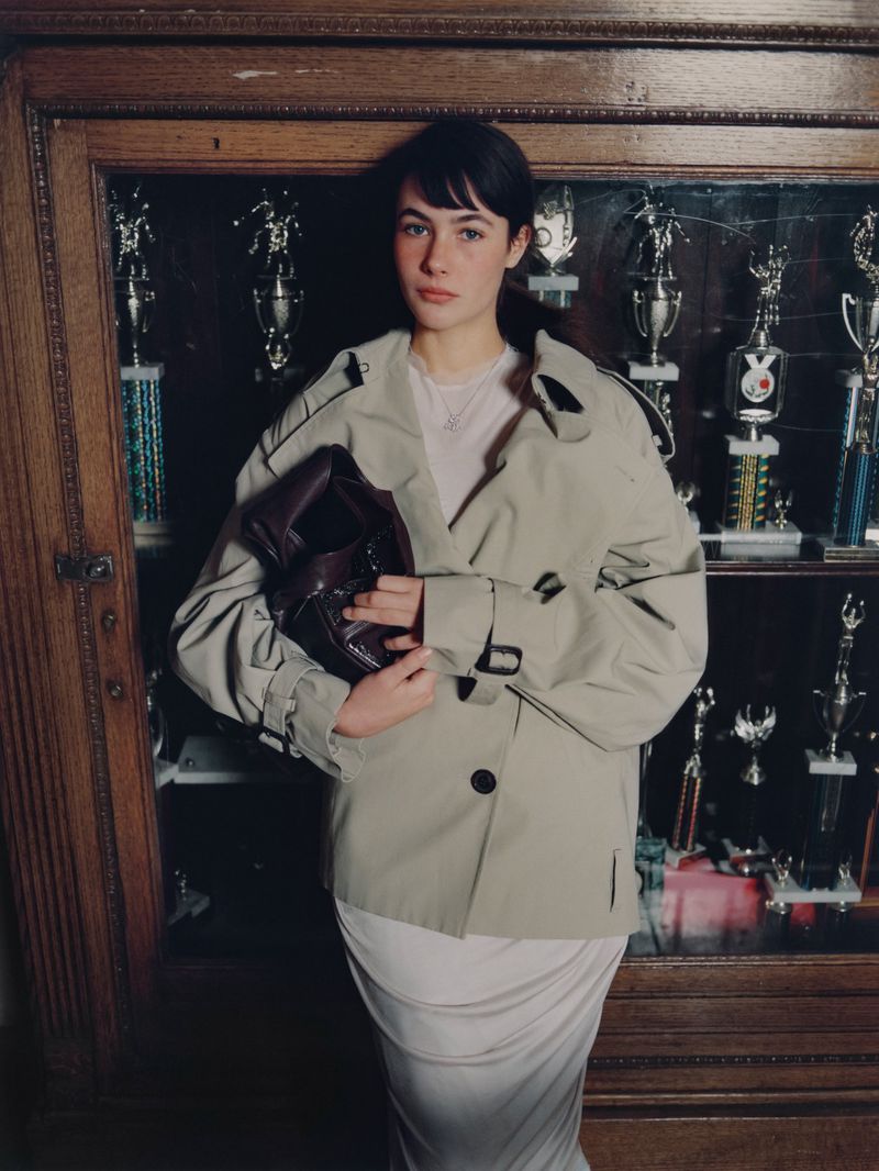 Woman poses in front of a wooden cabinet displaying trophies. She wears a trench coat and holds a leather bag.