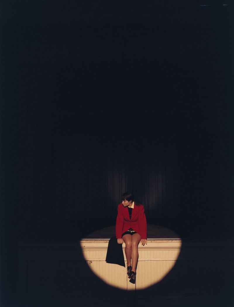 Woman in red jacket sits on ledge in spotlight against dark backdrop. Fashion shot with soft lighting and neutral color palette.