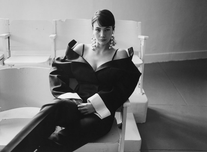 Monochrome portrait of an adult female in a dark suit with pearl earrings, sitting on a white chair in an indoor environment.