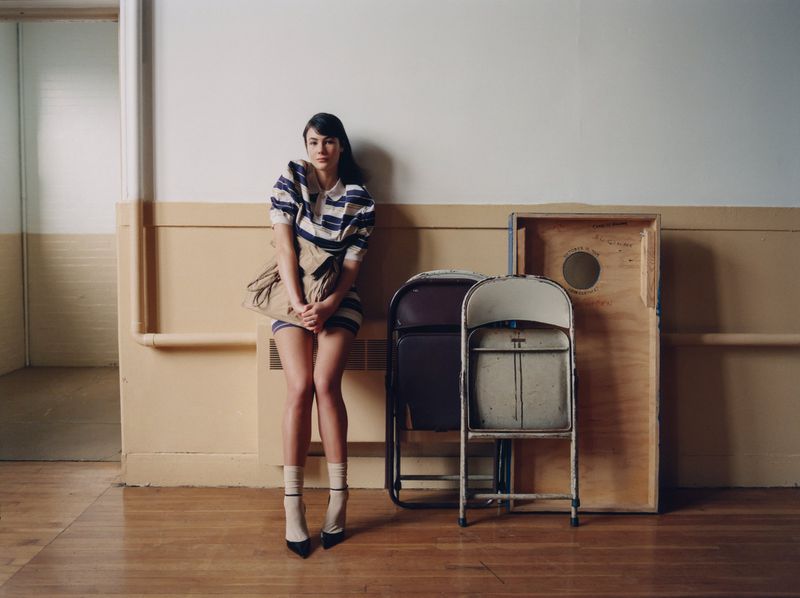 A woman with long dark hair in a blue and white striped dress poses next to folding chairs in an indoor location with neutral walls.