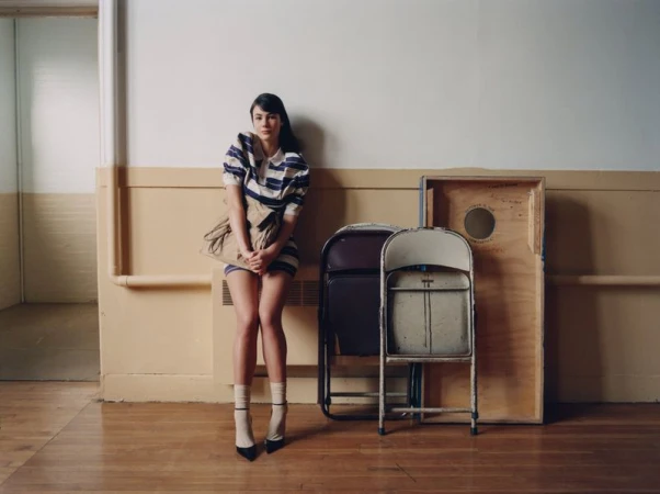 A woman with long dark hair in a blue and white striped dress poses next to folding chairs in an indoor location with neutral walls.