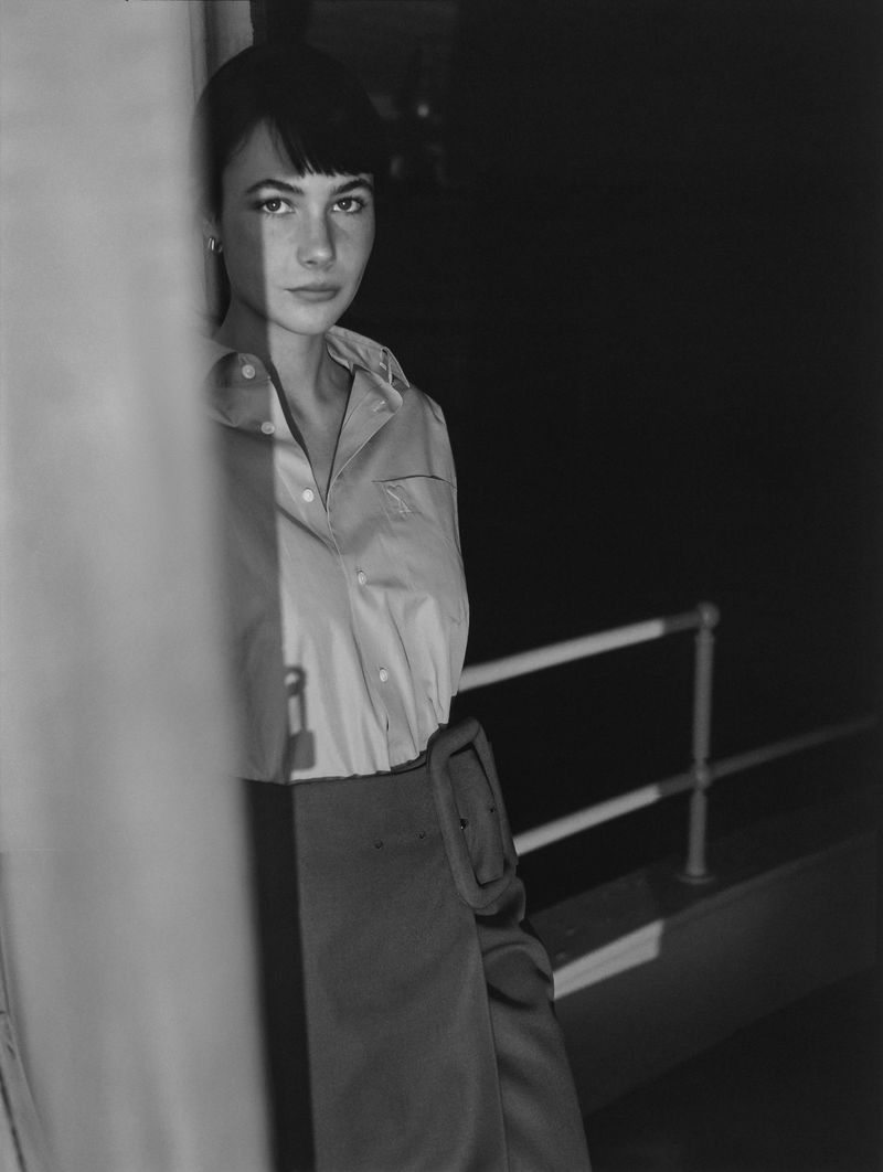 Monochrome portrait of an adult woman with short dark hair and bangs, wearing a collared shirt and dark skirt, looking at the camera.
