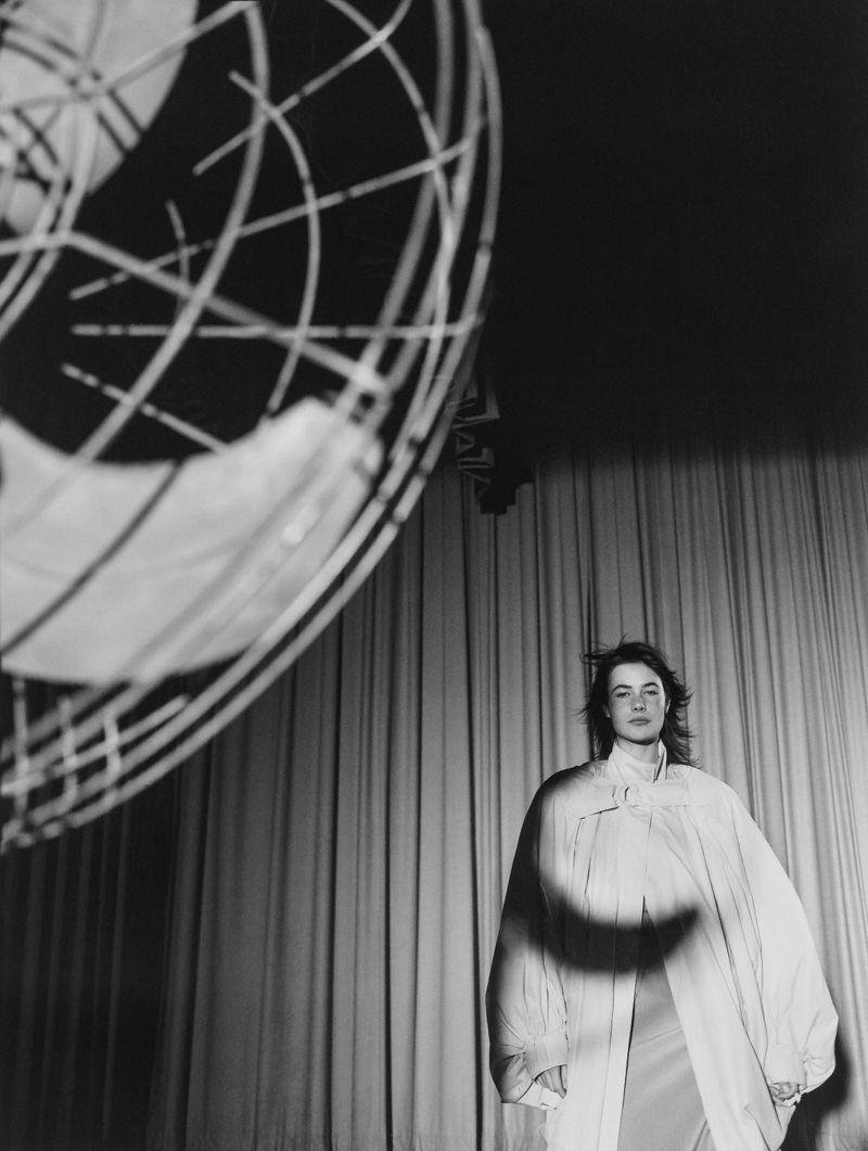Monochrome portrait of a woman in a coat standing in front of a curtain with a large spherical light fixture in the background.