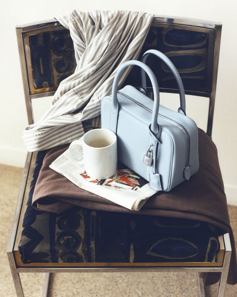 Light blue leather handbag on a metal chair with striped shirt, coffee cup, newspaper, and brown blanket, studio lighting.