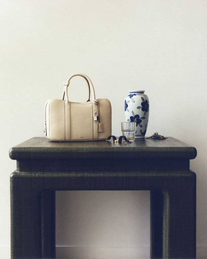 Beige leather handbag on a dark green table with a white vase, a clear glass, and some accessories, against a neutral backdrop.