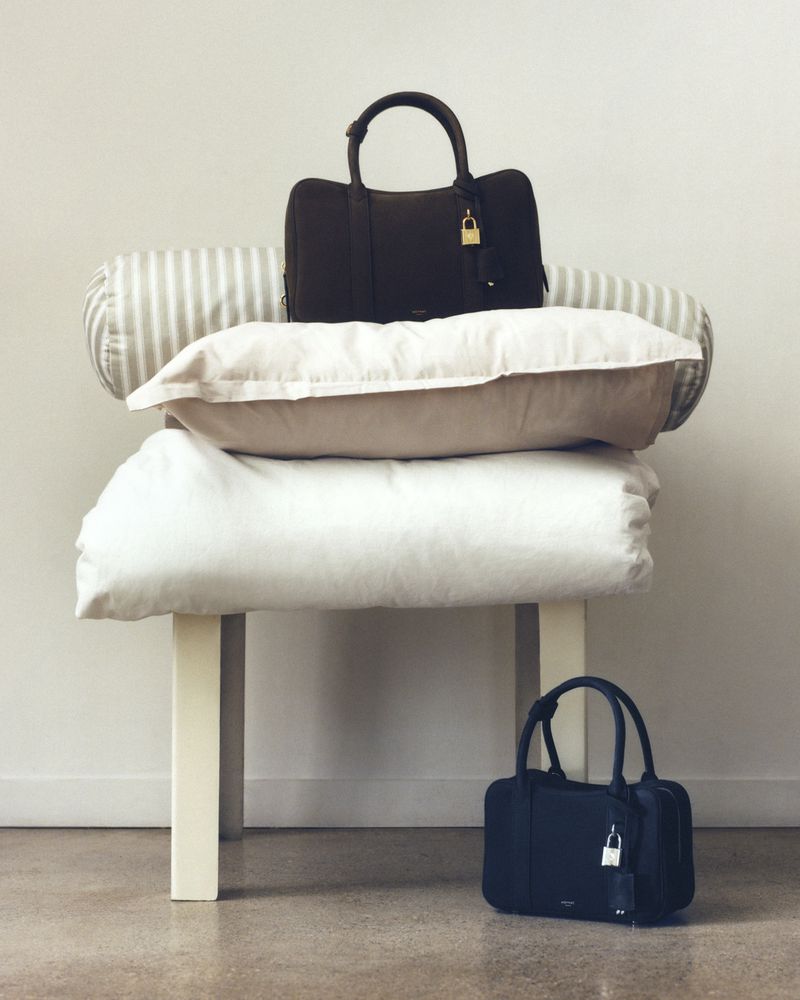 Two handbags are stacked on top of pillows and a bench in a neutral indoor setting, creating a visually appealing product arrangement.