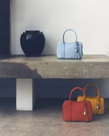Fashion still life featuring three handbags in blue, red, and yellow, arranged on a concrete surface with a black vase.