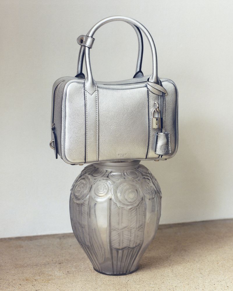 Silver metallic handbag with top handles and a padlock, resting on a decorative translucent glass vase with floral motifs.