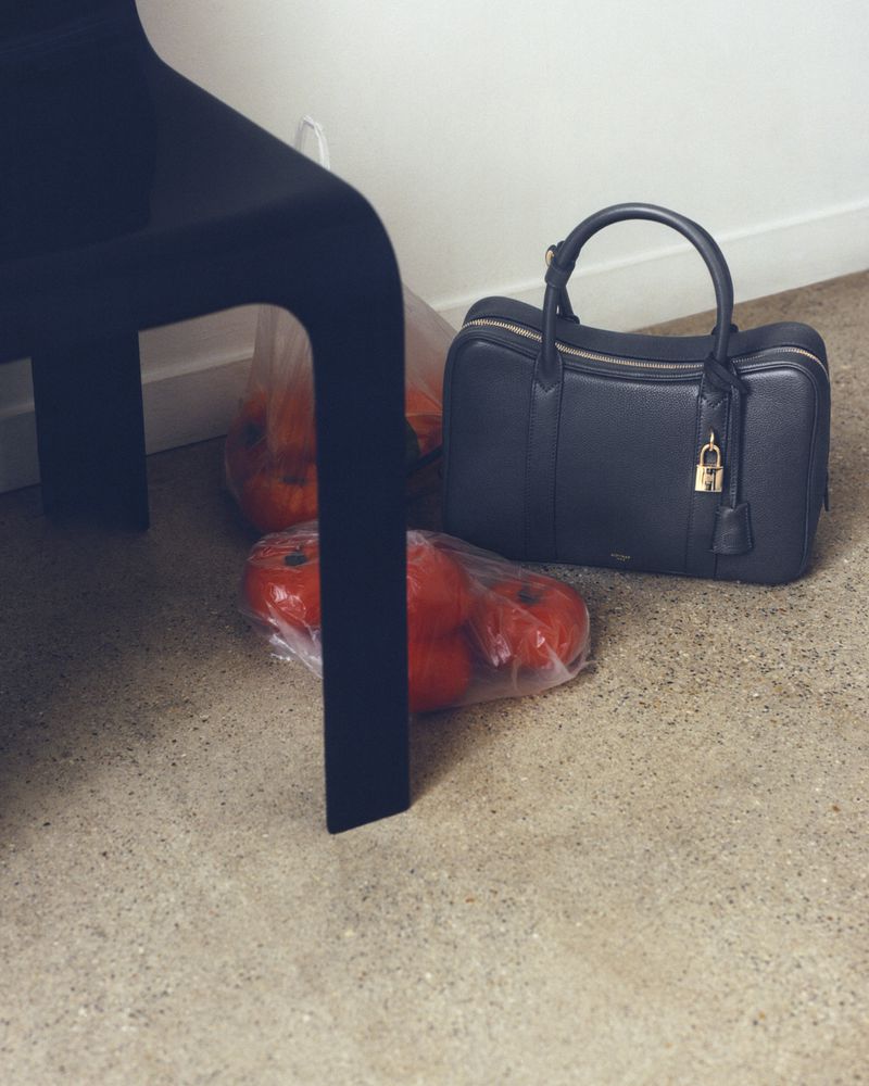 Black leather handbag next to bags of tomatoes on a beige floor, with a black chair in the background, showcasing home organization and food.