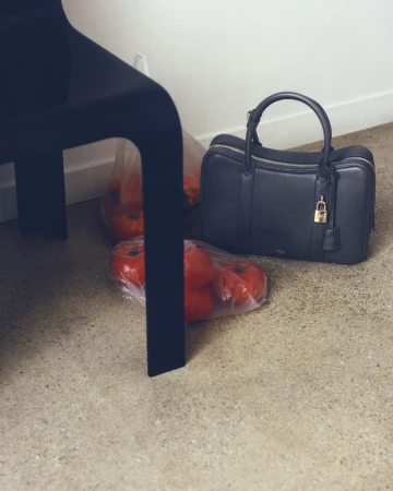 Black leather handbag next to bags of tomatoes on a beige floor, with a black chair in the background, showcasing home organization and food.