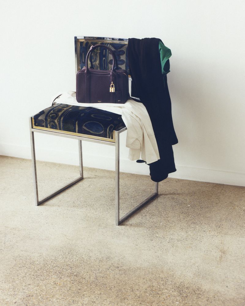 Studio shot of a silver bench with a dark blue, black and gold cushion, a dark purple handbag, and draped dark and light clothing.