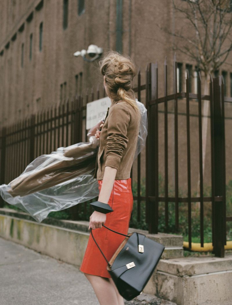 Woman in brown jacket and red skirt carrying a black handbag walks past a brown brick building on an overcast day.