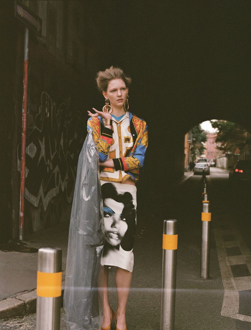 Fashion model in a patterned jacket and skirt with a face graphic, standing on a street with bollards, with a dark archway behind.