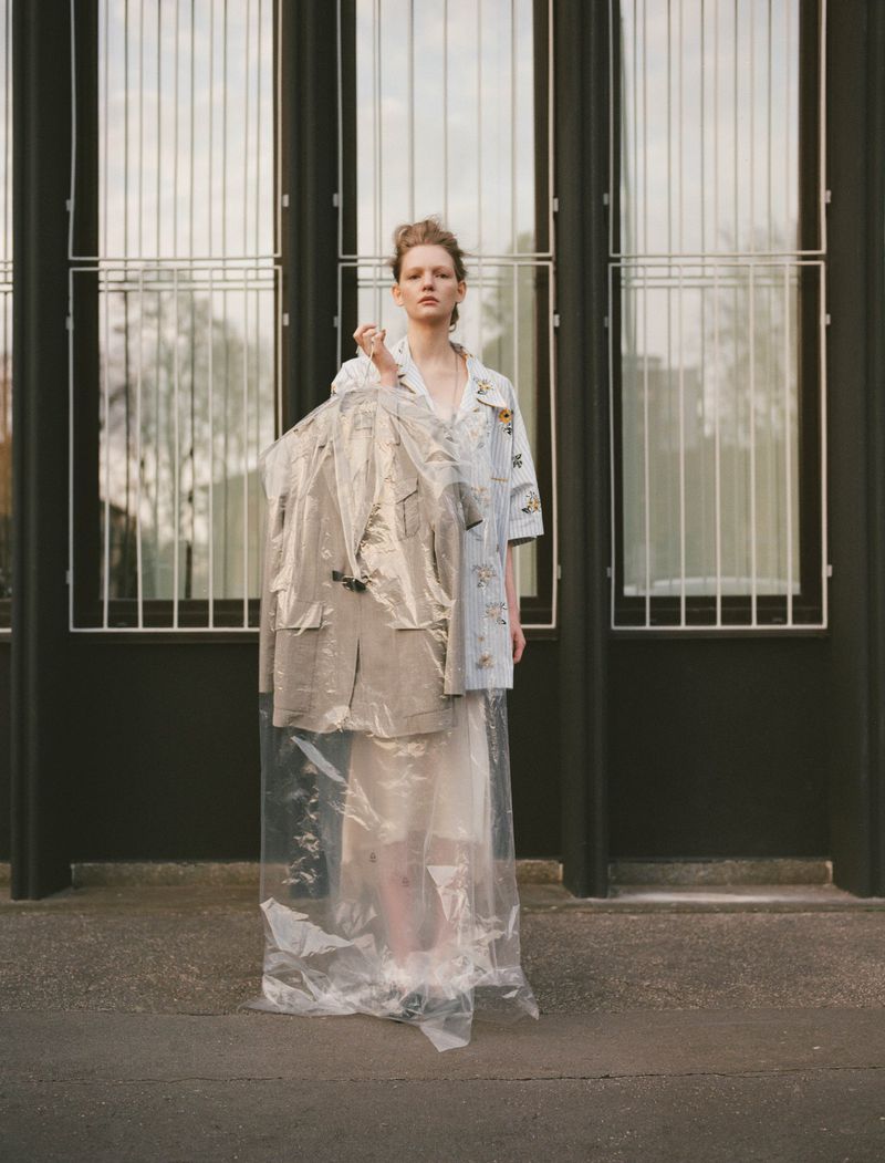 A fair-skinned young adult woman stands in front of a building, holding a suit in a plastic garment bag, wearing a patterned shirt and skirt.