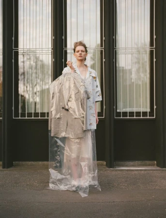 A fair-skinned young adult woman stands in front of a building, holding a suit in a plastic garment bag, wearing a patterned shirt and skirt.
