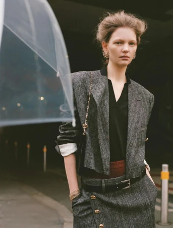Fashion editorial shot of a stylish young woman with an umbrella. She's wearing a grey tweed jacket, skirt and gold jewelry, in a dark setting.