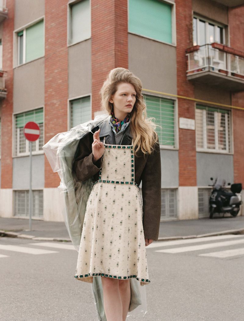 A woman poses on a city street wearing a patterned dress, a brown jacket, and a clear plastic coat with buildings in the background.