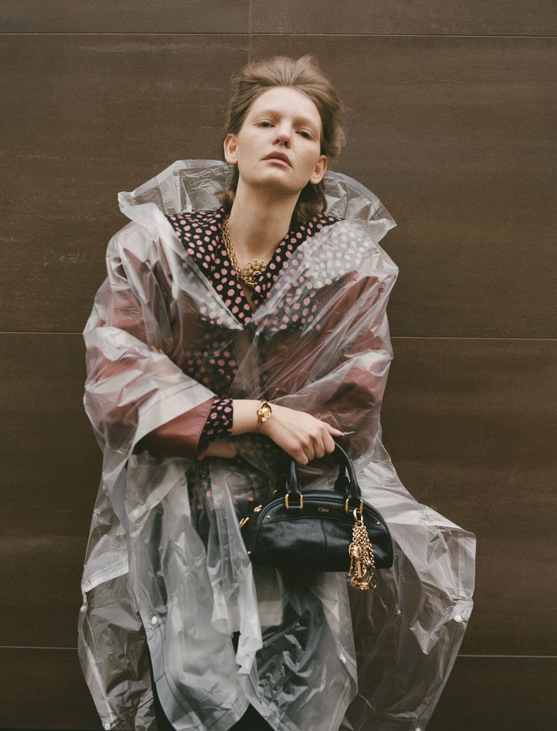 Blonde woman in a translucent plastic raincoat and polka dot blouse, carrying a black purse against a brown tiled wall.